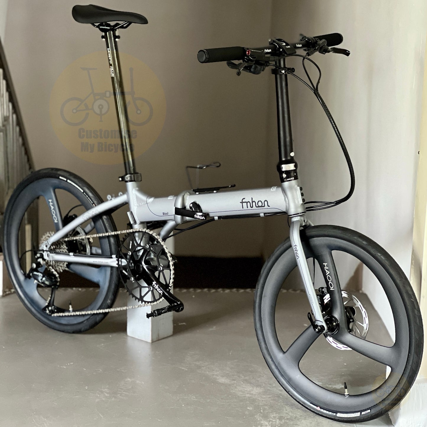 A folding electric bicycle with a silver frame and black components, photographed indoors against a beige wall. The bike has a visible 'fnhan' logo on the main tube and 'Borama' branding on the wheel spokes.